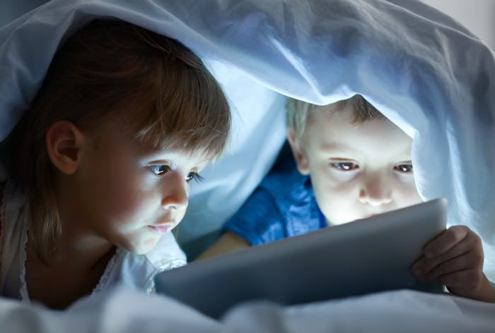 Cute little children lying under blanket with tablet computer