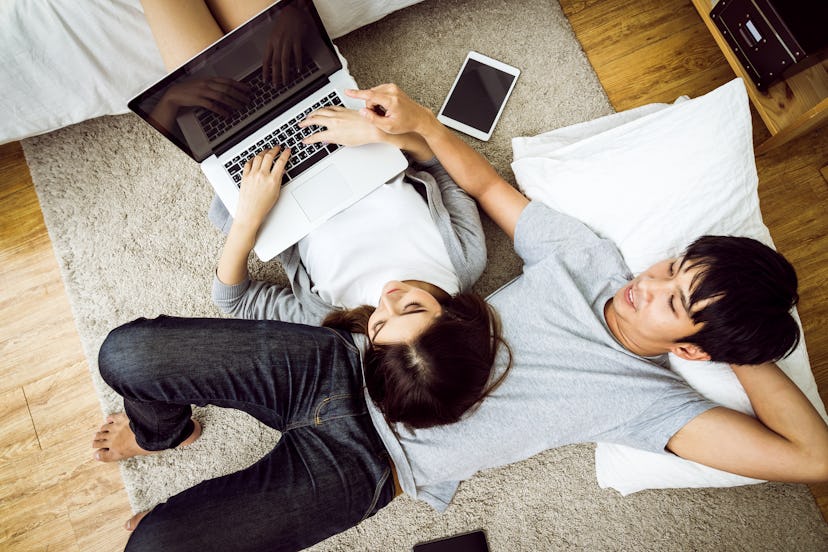 Happy asian couple using or looking at laptop computer with smile lying together on wooden floor wat...