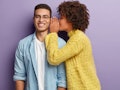 Afro American female whispers secret to boyfriend who has cheerful expression, gossip together, wear...
