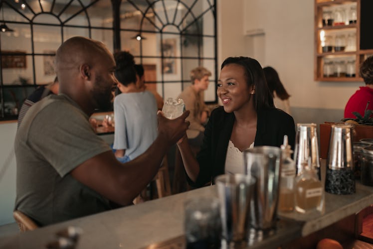 Smiling young African American couple sitting at the counter of a bar toasting with drinks while on ...