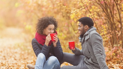 Cozy fall. Afro couple drinking hot coffee, having picnic in forest, sitting on plaid