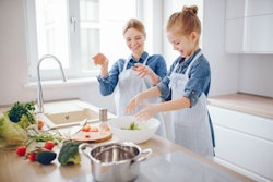 A young and beautiful mother in a blue shirt and apron is preparing a fresh vegetable salad at home ...