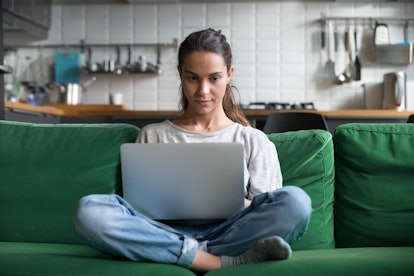 A young woman sits on her green couch and looks at her laptop.