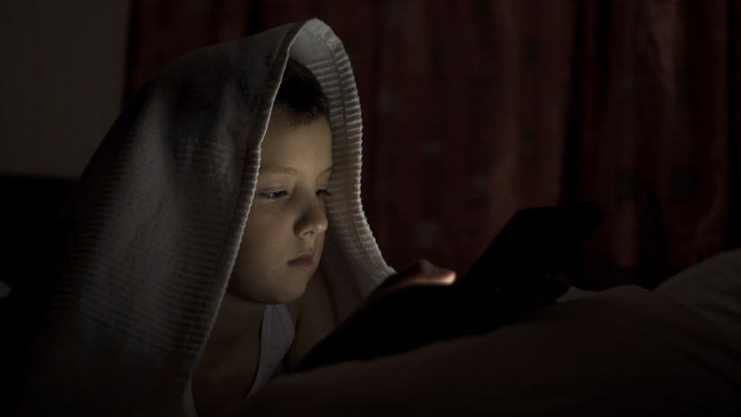A happy boy lies in bed under a blanket and plays on a tablet in a game in the dark. The face of the...