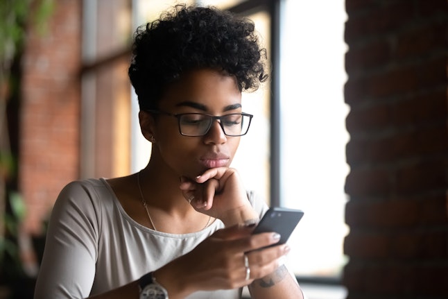 Serious black woman wearing glasses holding phone browse internet reading media news downloading new application or game. Student typing message using messenger or customer buying goods online concept