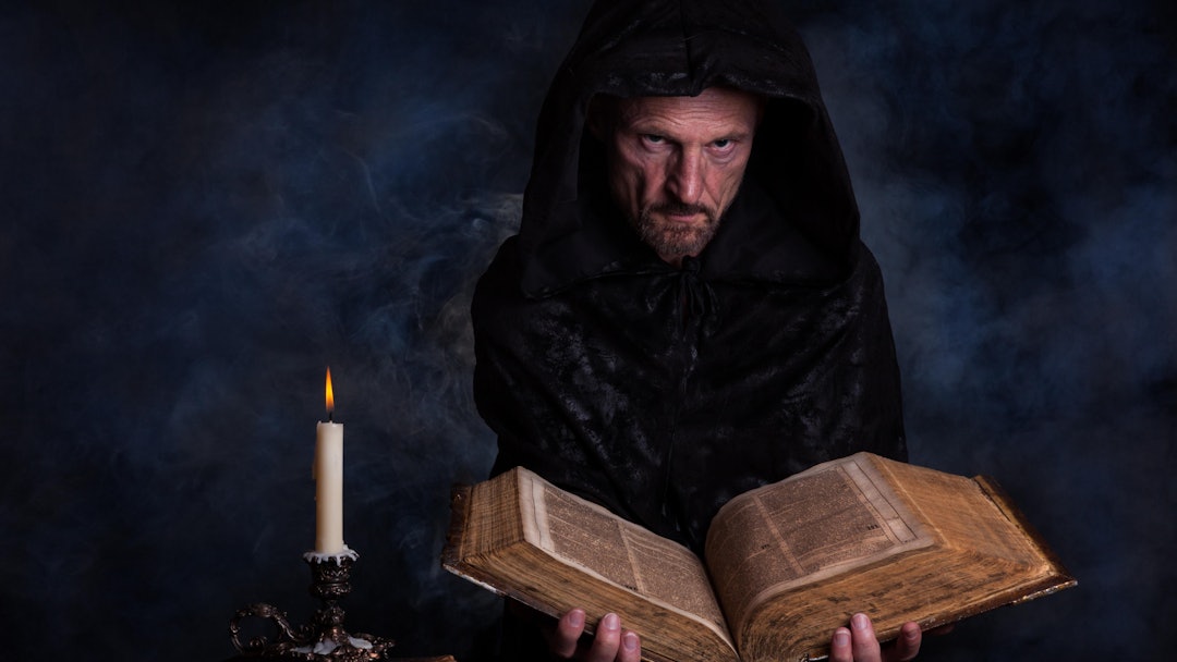 MODEL RELEASED Man, with a black hooded coat, holding an old book in his hands, in front a burning c...