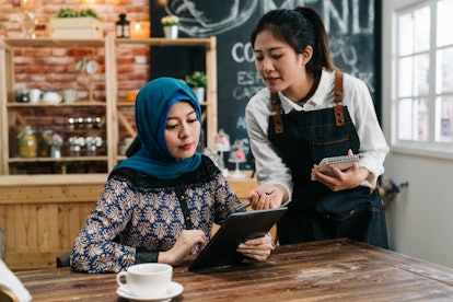 Young Asian waitress advising islam woman client to choose new snack from menu on mobile pad in rest...