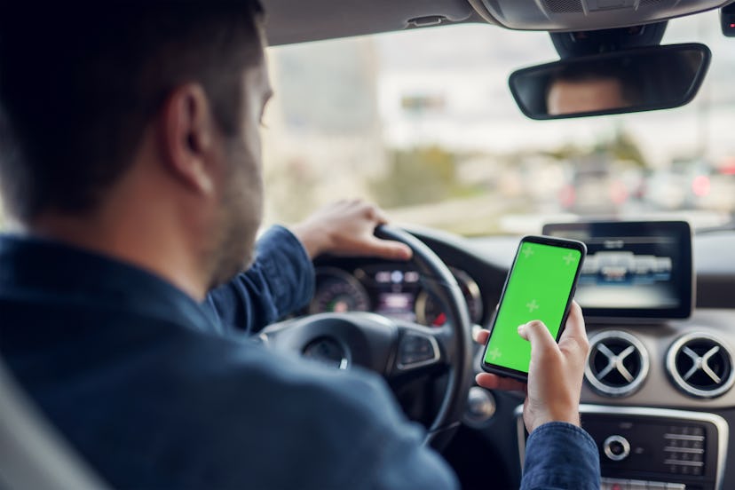 Photo from back of man with phone with green screen in his hands at wheel of car