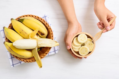 Hand holding fork for eating sliced banana in a bowl and peeled banana in a basket, top view