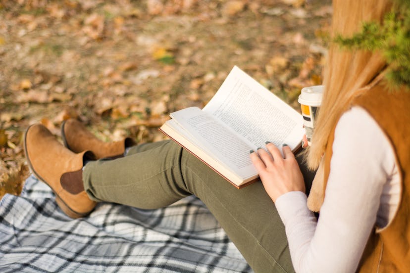 Young girl reading and holding book on legs at retro blanket at park on autumn day. Fall leaf blurre...