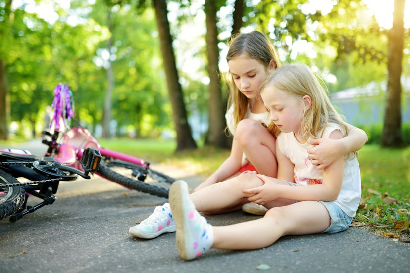 Adorable girl comforting her little sister after she fell off her bike at summer park. Child getting...