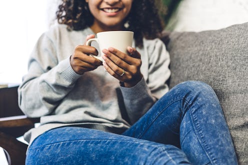 Young african american black woman relaxing drinking cup of hot coffee or tea on couch at home