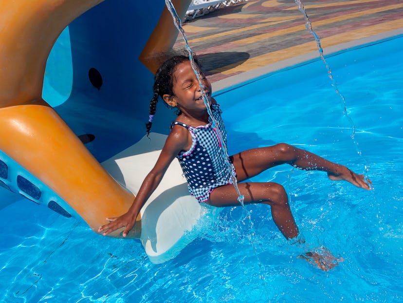 Mixed ethnicity little smiling girl coming down on the chute into the water. On the slide in aqua pa...