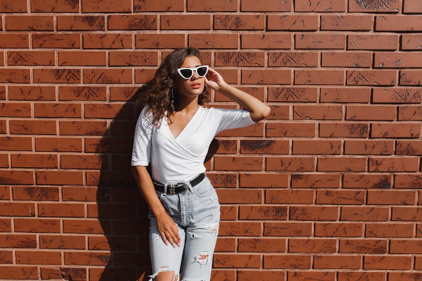 Attarctive elegant young woman hipster in a white sunglasses in a summer denim pants and white t-shi...
