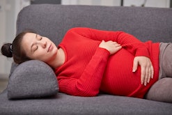 Photo of sleepy tired pregnant woman takes nap, keeps hands on belly, wears red warm sweater, enjoys...