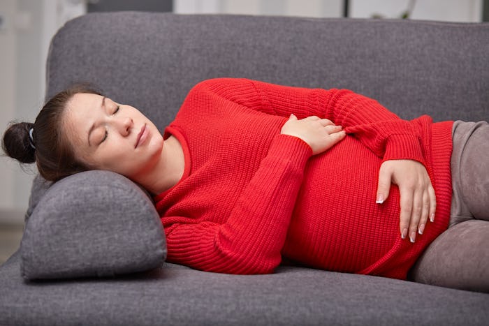 Photo of sleepy tired pregnant woman takes nap, keeps hands on belly, wears red warm sweater, enjoys...