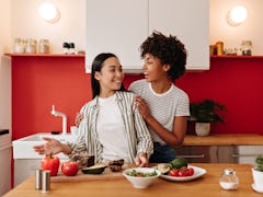 Girl hugs friend by shoulders in kitchen. Dark-haired women make salad