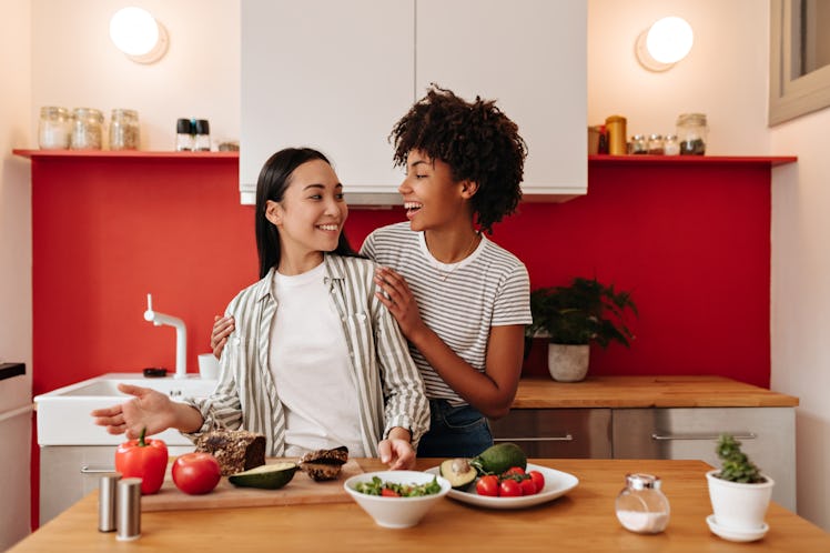 Girl hugs friend by shoulders in kitchen. Dark-haired women make salad