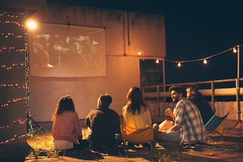 Group of young friends watching a movie on a building rooftop terrace, eating popcorn, drinking beer...