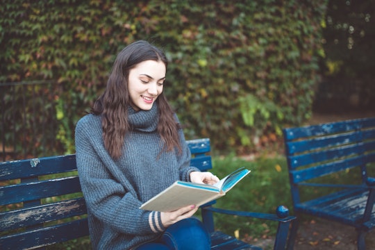 Young woman in a sweater reading a book, sitting on the bench in the park. Autumn time.