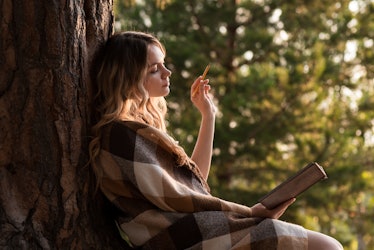 A girl sits leaning on a tree writing in a notebook outdoors.  Checkered plaid on shoulders. Thought...