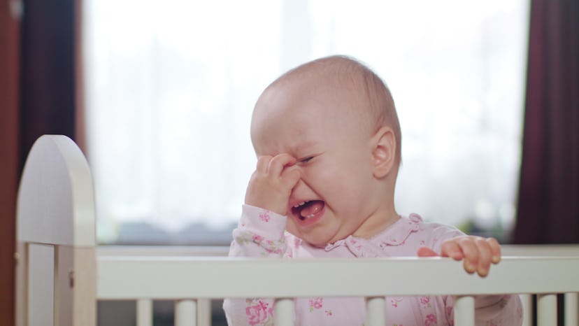 Baby's standing in a white crib at home and crying. Medium shot.