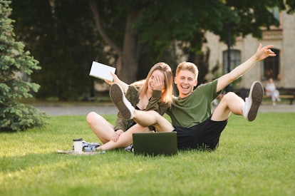 Two happy joyful students fooling around sitting on college lawn with books, laptop and take away co...