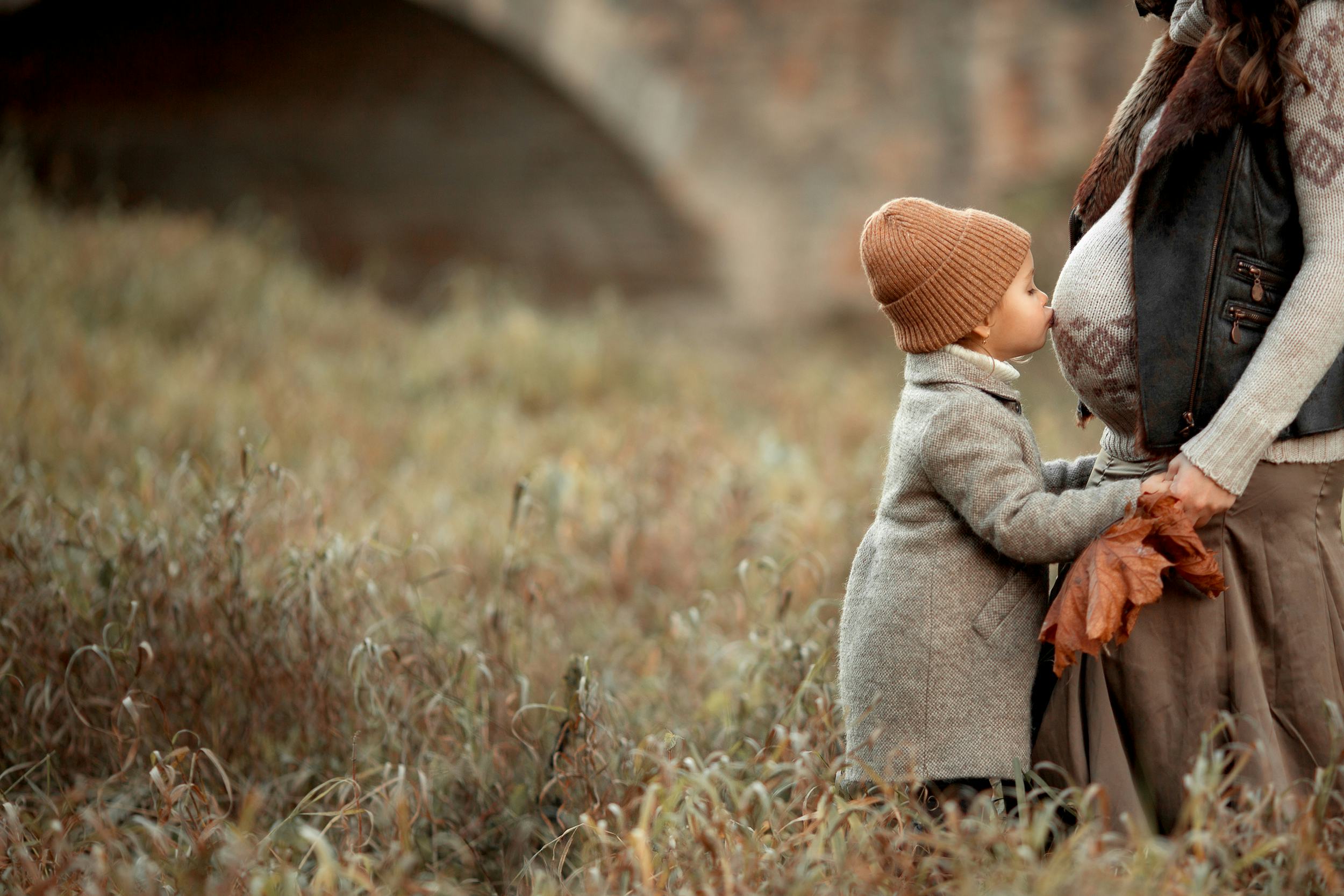 pretty little girl in brown hat and coat standing in the meadow in the park and kiss stomach of her ...