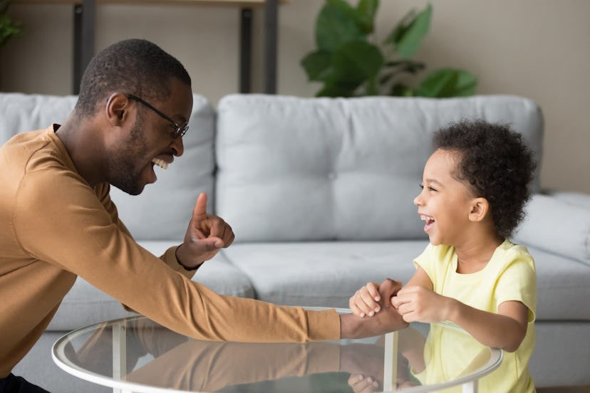 Laughing black father and biracial cheerful son having fun on weekend try to play explain arm wrestl...