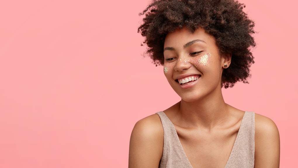Studio shot of happy young African American female has glitter on cheeks, smiles happily with shy expression, has dark skin, stands against pink backgrounf with free space for your promotion