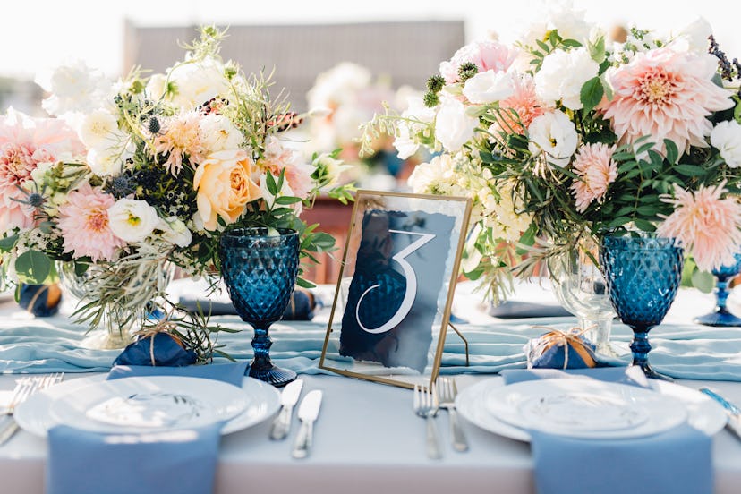 Long dinner tables covered with white cloth, served with porcelain and blue glasses and rich decorat...