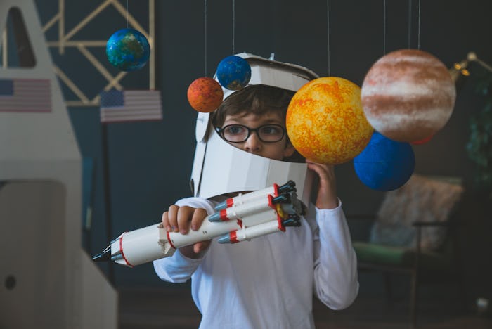 Cute little boy wearing cardboard astronaut helmet flying toy rocket through planets, cardboard spac...