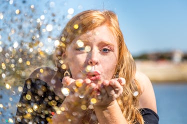 A woman with red hair blows glitter out of her hands on a sunny day at the beach.