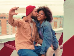 Kiss and selfie. Loving young man kissing his smiling happy girlfriend while sitting on the roof wit...