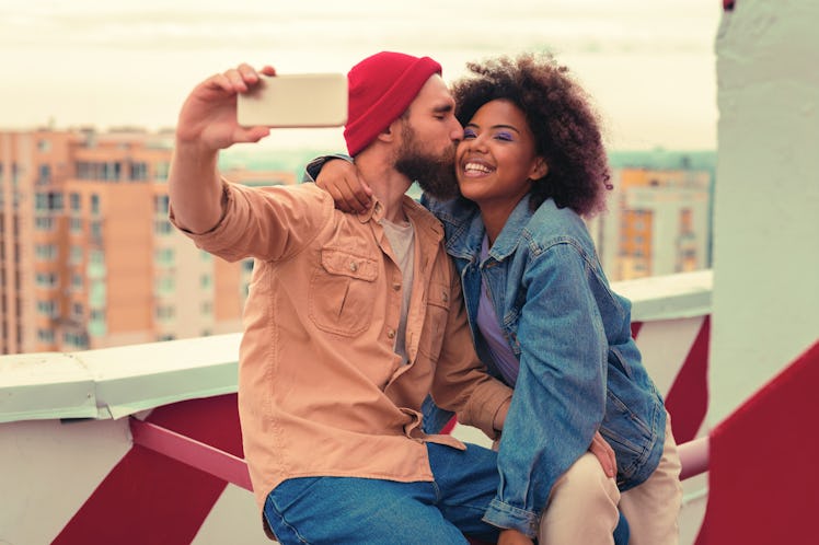 Kiss and selfie. Loving young man kissing his smiling happy girlfriend while sitting on the roof wit...