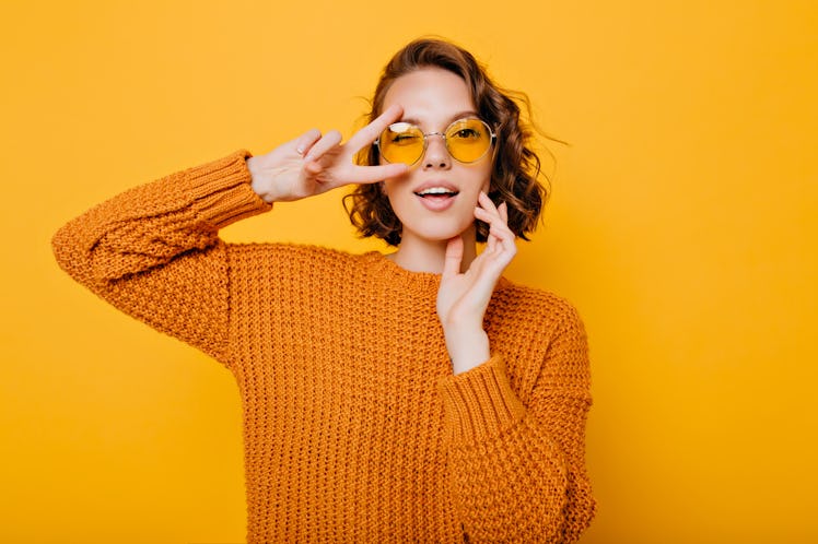 Close-up portrait of charming young woman with gently smile posing with peace sign in trendy glasses...