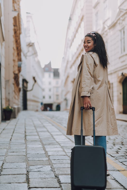 A happy woman rolls her suitcase down a city street in Europe while on vacation.