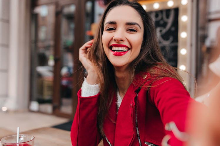A happy brunette woman in a red outfit takes a selfie while sitting outside a café on Valentine's Da...