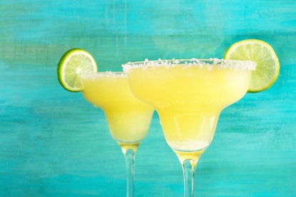 A closeup of two lemon Margarita cocktails with wedges of lime on a vibrant turquoise background wit...