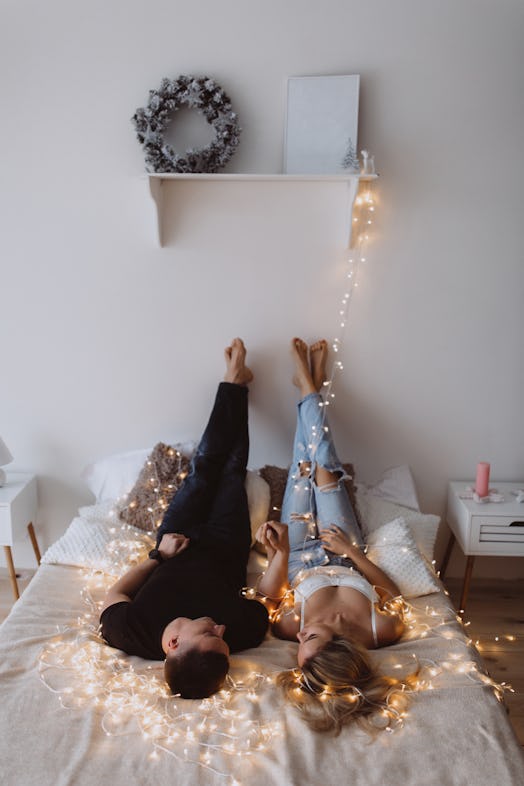 A young couple looks at each other and lays in a bed covered in string lights.