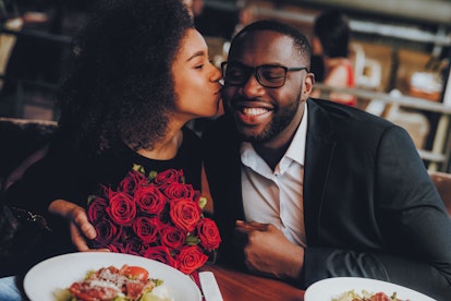 A woman holding a bouquet of red roses kisses her boyfriend on the cheek in a restaurant.