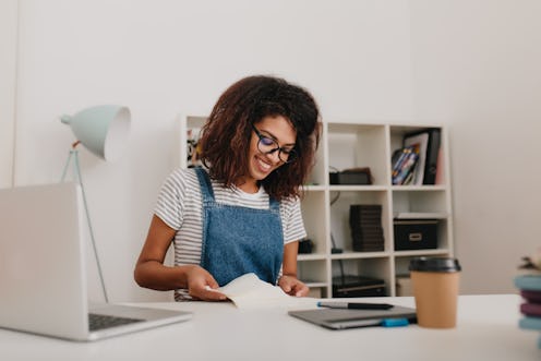 Graceful girl with curly brown hair reading documents with pretty smile sitting in her office. Indoo...