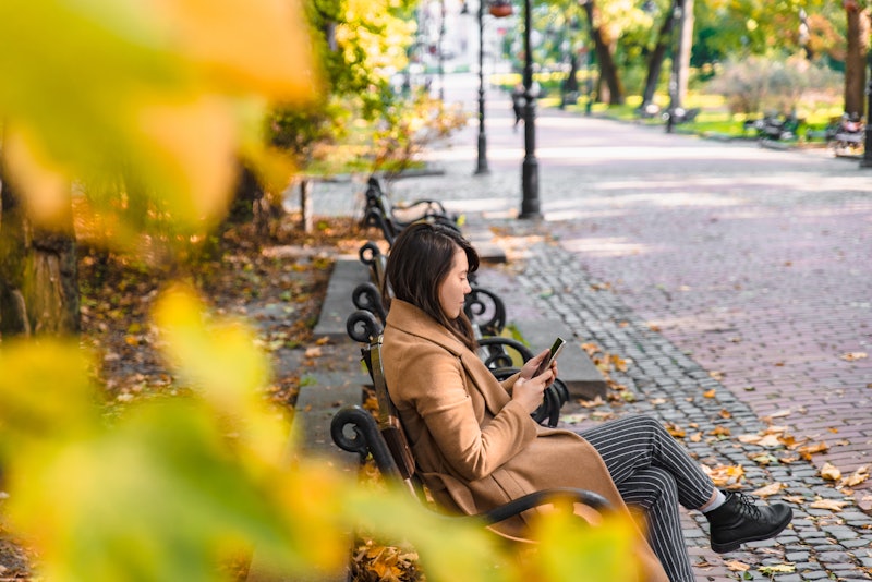 young adult fashion woman sitting at city park bench in autumn fall season sunny day