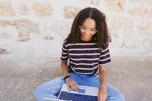 A portrait of happy smiling young black woman with curly hair wearing glasses, jeans and a striped t...