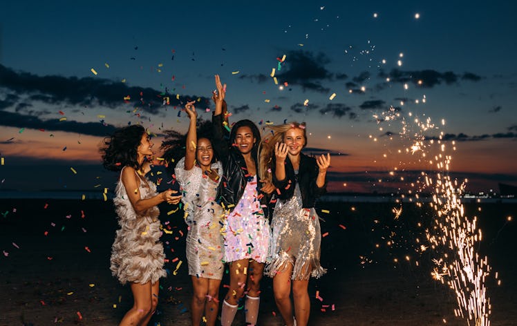A group of friends celebrate a birthday on the beach with fireworks, confetti, and party dresses.