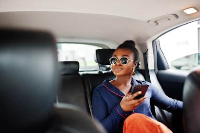 A chic woman sits in the back of a car and drives around a city with her phone in her hand.