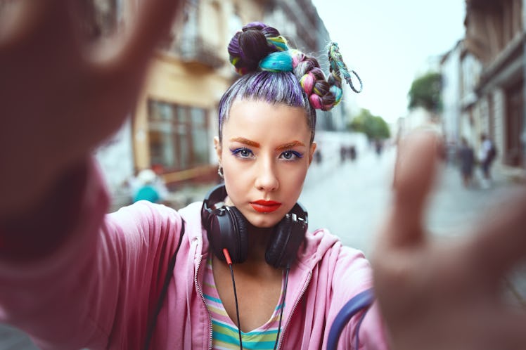 Cool funky young girl with headphones and crazy hair enjoy power of music taking selfie on street – ...