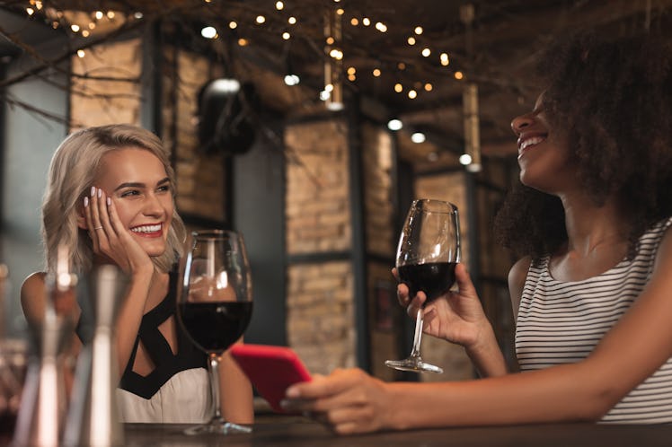 Amusing stories. Cheerful young women sitting at the bar counter, drinking wine and laughing heartil...