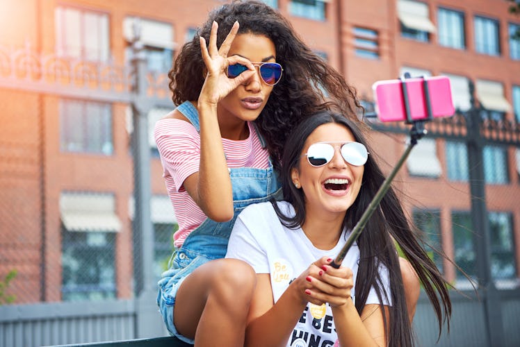 Two happy girls wearing cool sunglasses sit on a bench, laugh, and pose for a selfie on a summer day...