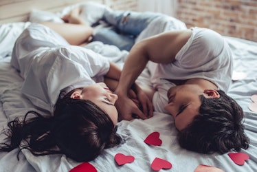 Beautiful young couple in bedroom is lying on bed with red hearts nearby. Enjoying spending time tog...
